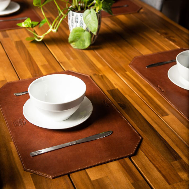 Beveled edge personalized fine leather placemat in brown, set on a wooden table with white dishware.