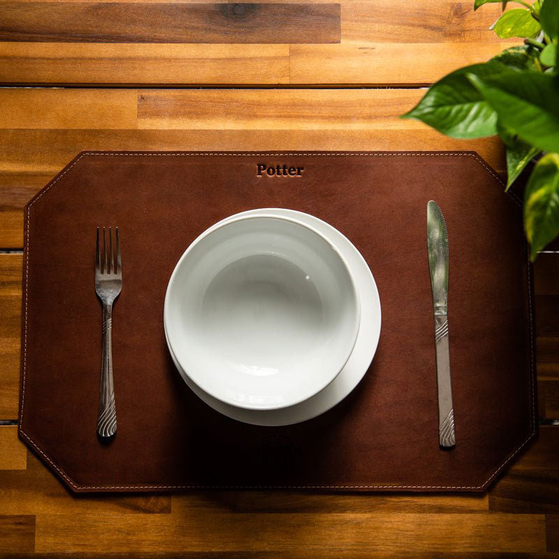 Beveled edge personalized fine leather placemat in brown with a bowl, plate, fork, and knife set on a wooden table.