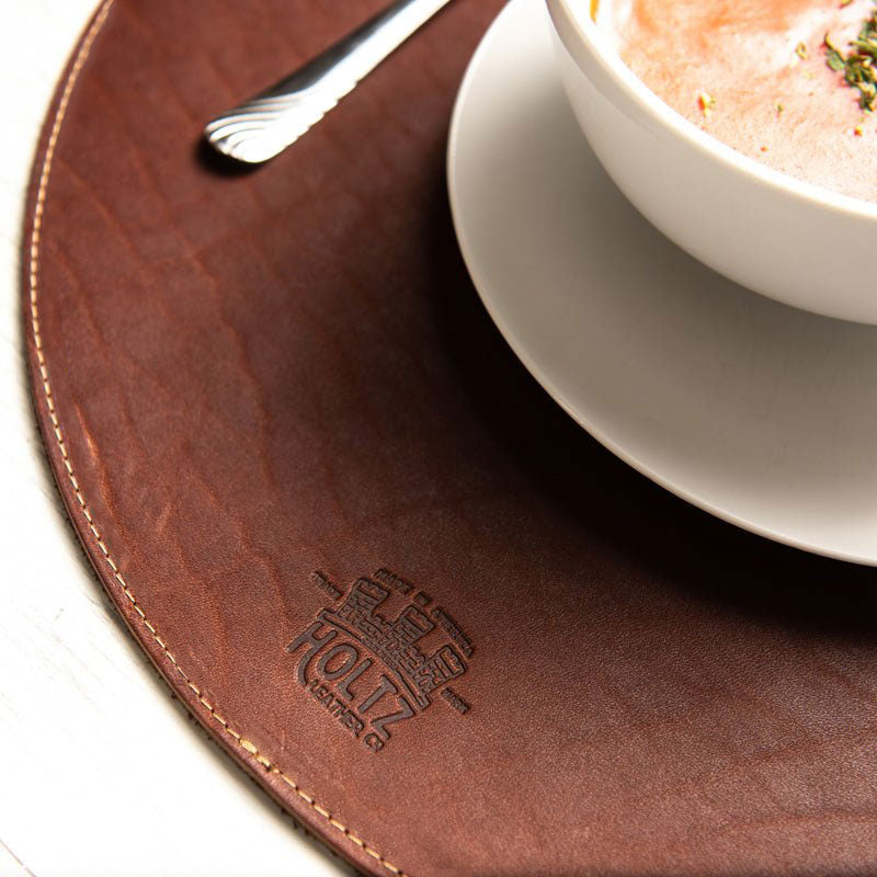 Circle personalized fine leather placemat in rich brown with embossed logo, featuring a bowl and spoon on top.