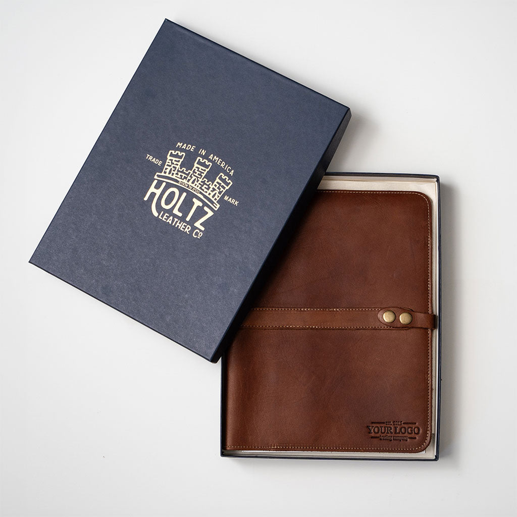 A brown leather folio-style wallet or notebook cover displayed next to its dark navy blue presentation box on a light gray background. The box features gold foil branding for 'Holtz Leather Co.