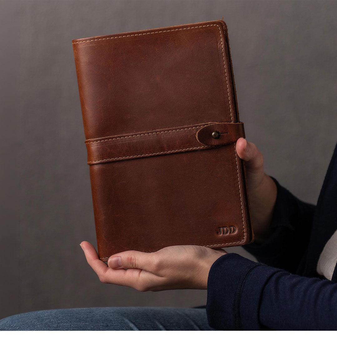 A person holds a brown leather planner or notebook cover with a strap closure. The cover has a debossed 'JDD' logo in the bottom right corner and is shown against a dark gray background.