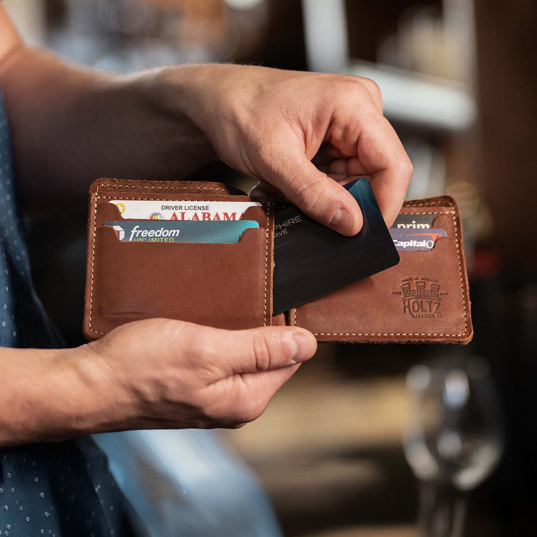 A close-up of a person holding an open brown leather wallet with card slots and embossed branding, inserting a black card. The wallet has visible cards inside and is photographed in a softly blurred indoor setting.