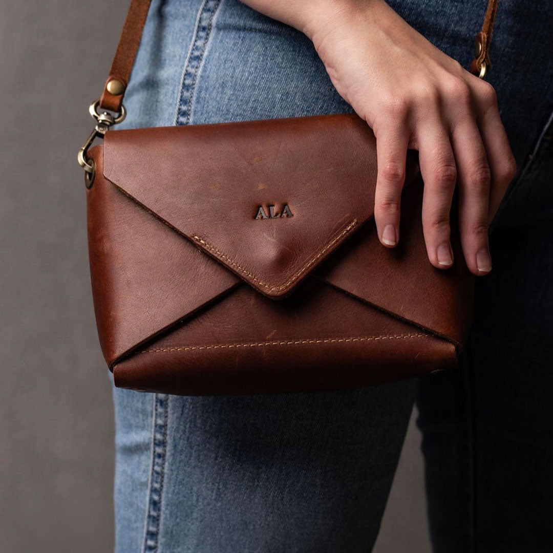 A brown leather envelope-style shoulder bag with a flap closure and gold-tone hardware, shown being held by a person wearing blue jeans. The bag has 'ALA' embossed on the front flap.