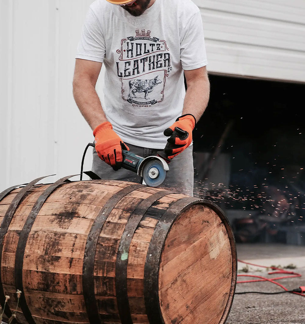 A person wearing gloves and a hard hat uses a power tool to cut or sand a wooden barrel, with sparks flying beside an open garage door.