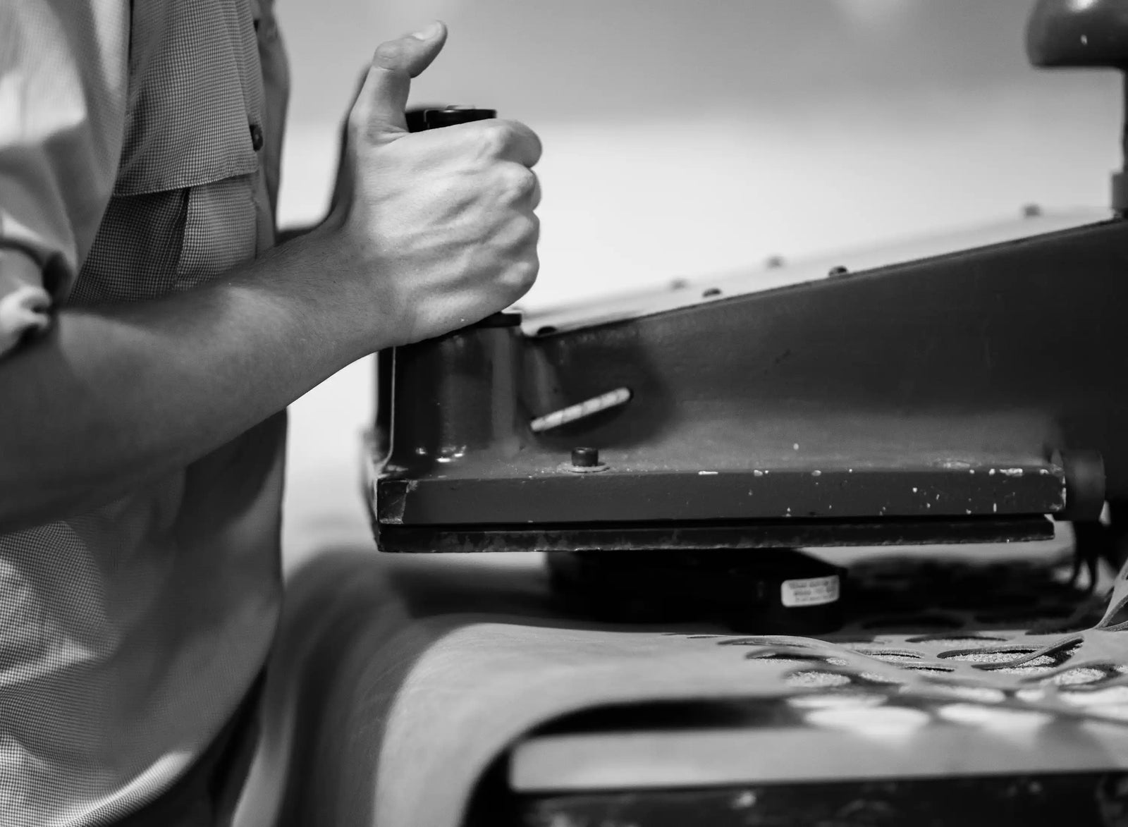 Black-and-white close-up of a hand operating an industrial sewing machine, with leather beneath it.
