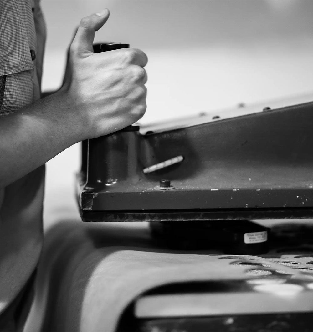 Black-and-white close-up of a hand operating an industrial sewing machine, with leather beneath it.