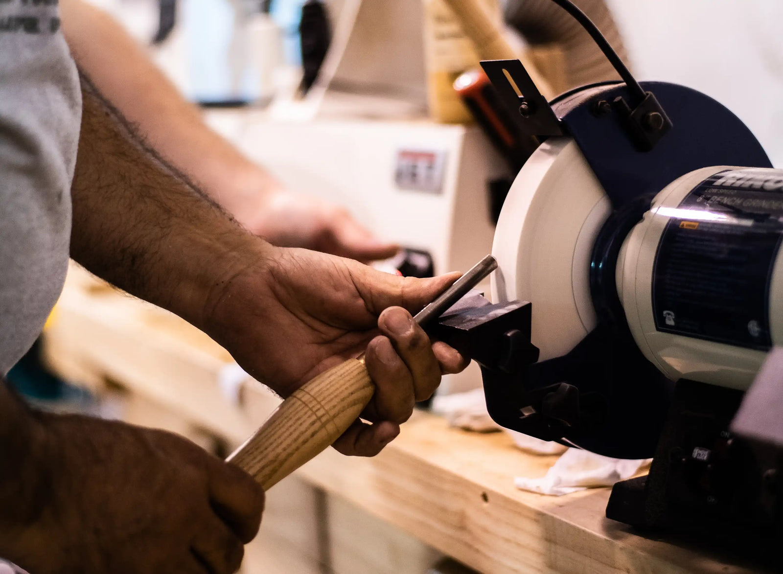 Close-up of a craftsperson sharpening a metal tool against a rotating workshop grinder, with wood shavings and equipment in the background.