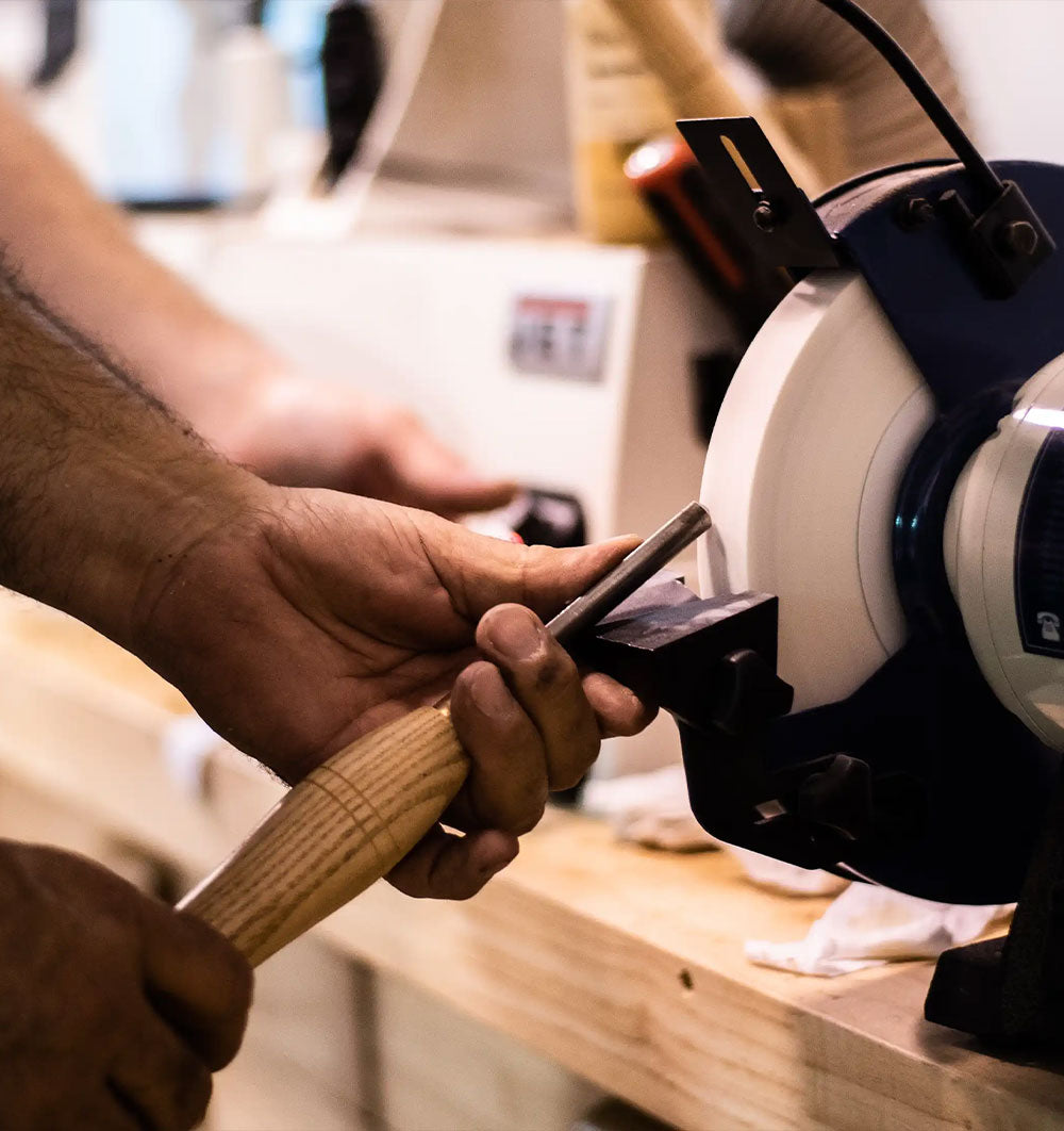 A close-up action shot of a person using a handheld sharpening stone on a metal rod, with a woodworking bench in the background.