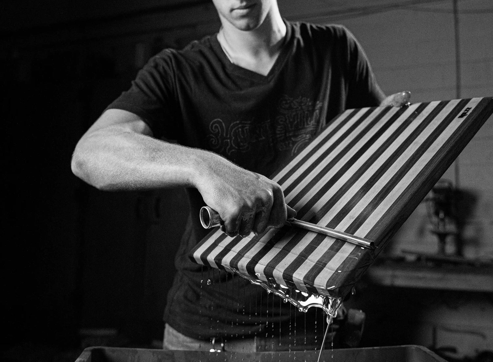 Black-and-white photo of a man tilting a striped wooden board as water drips into a basin, creating a moody, hands-on workshop scene.