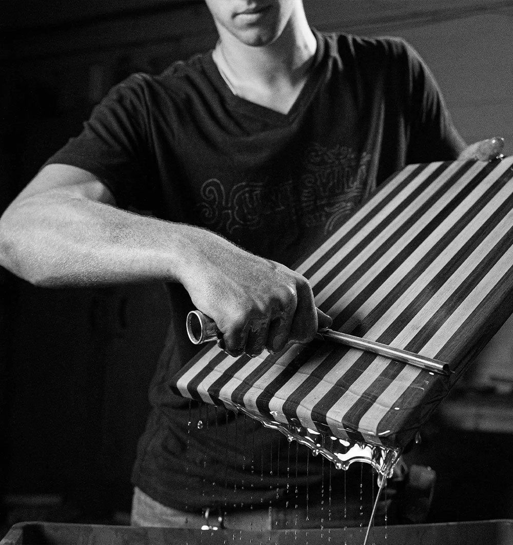 Black-and-white photo of a man tilting a striped wooden board as water drips into a basin, creating a moody, hands-on workshop scene.