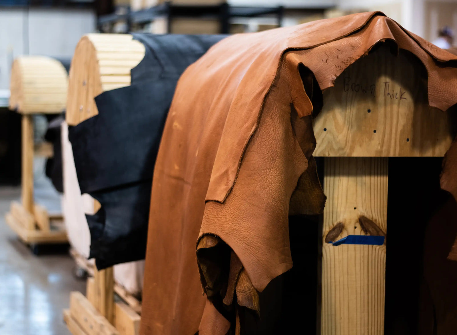 Leather hides drape over wooden forms in a workshop, showcasing warm brown and dark materials in a rustic, handcrafted setting.