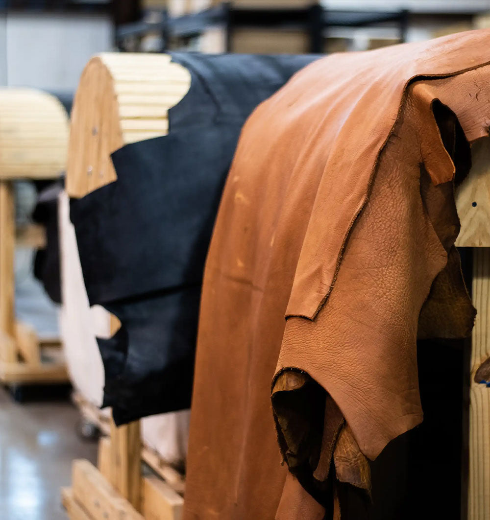 Leather hides drape over wooden forms in a workshop, showcasing warm brown and dark materials in a rustic, handcrafted setting.