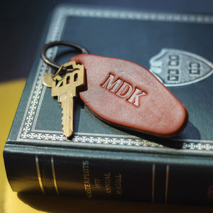 A close-up of a leather keychain with the initials 'MDK' embossed on it, attached to a brass key and resting on top of a dark decorative book.