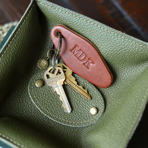 An olive green textured leather key holder pouch containing a brown leather key tag embossed with 'MDK' and two keys, shown in a close-up shot.