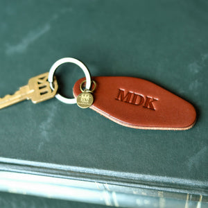 A close-up of a brown leather keychain with the initials 'MDK' embossed on it, attached to a silver split ring and a gold-toned key. The keychain rests on a dark green textured surface.
