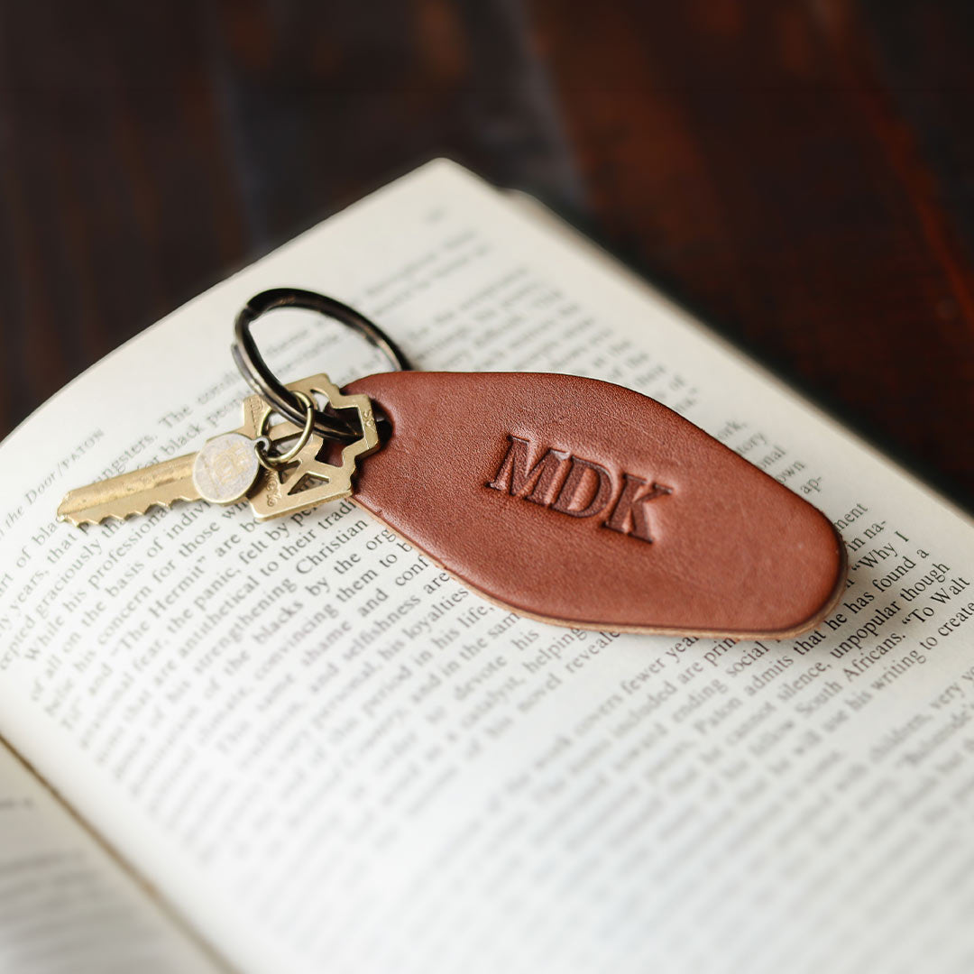 A brown leather keychain with a monogrammed 'MDK' tag and brass keys, resting on an open book over a dark wooden surface.